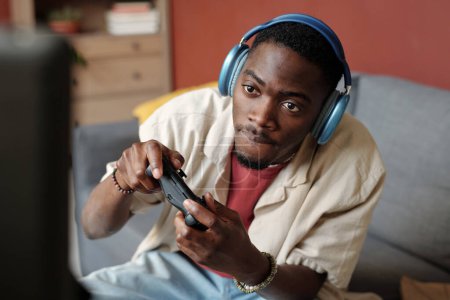 Téléchargez les photos : Joueur masculin tendu avec des contrôleurs dans les mains regardant l'écran de l'ordinateur tout en jouant au jeu vidéo à loisir dans l'environnement de la maison - en image libre de droit