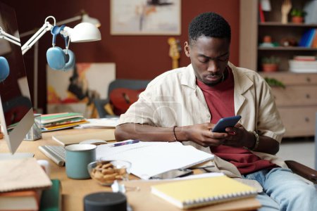 Téléchargez les photos : Jeune freelance masculin regardant l'écran du smartphone tout en étant assis sur le lieu de travail avec des papiers et des blocs-notes et en organisant le travail - en image libre de droit