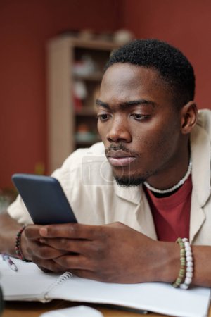 Foto de Joven estudiante masculino serio mirando la pantalla del teléfono inteligente mientras está sentado junto a la mesa con copybook abierto delante de la cámara y viendo video - Imagen libre de derechos