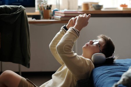 Photo for Side view of youthful boy in headphones sitting against bed and looking at screen of mobile gadget connected to gamepad during video gaming - Royalty Free Image