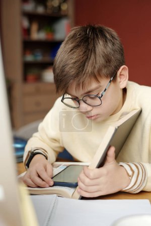 Photo for Serious schoolkid in eyeglasses looking at screen of smartphone on page of open textbook while preparing home assignment - Royalty Free Image