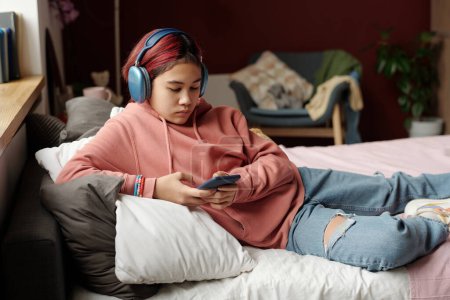 Téléchargez les photos : Adolescent mignon en casual et écouteurs se détendre sur le lit et en regardant l'écran du smartphone tout en faisant défiler la liste de lecture - en image libre de droit