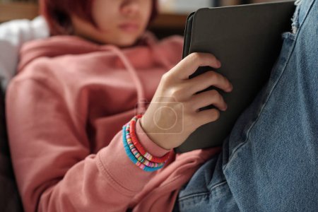 Téléchargez les photos : Main d'adolescente méconnaissable en sweat à capuche rose tenant la tablette tout en étant assis devant la caméra sur le lit et faisant des devoirs - en image libre de droit