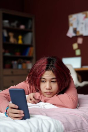 Photo for Teenage girl scrolling through fresh online posts in social networks or watching video while relaxing on bed after school - Royalty Free Image