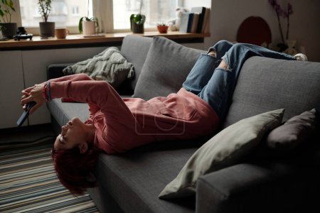 Photo for Pretty teenage girl in casualwear lying on comfortable couch in living room and looking at smartphone screen while watching online video - Royalty Free Image