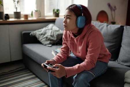 Photo for Serious teenage girl in headphones sitting on couch in front of camera and pressing buttons of joystick while playing video game - Royalty Free Image