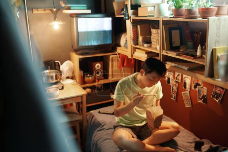 Foto de Joven mujer asiática en ropa de casa sentada en la cama por el viejo televisor y estantes de madera y comiendo wok chino de cartón lonchera - Imagen libre de derechos