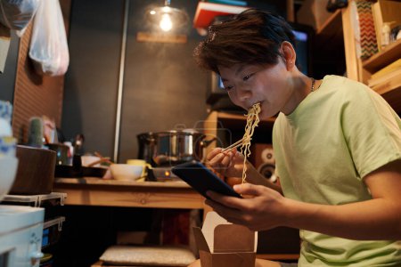 Foto de Mujer asiática joven en camiseta mensajes de texto o ver vídeo en línea en el teléfono inteligente y comer wok chino de cartón lonchera - Imagen libre de derechos