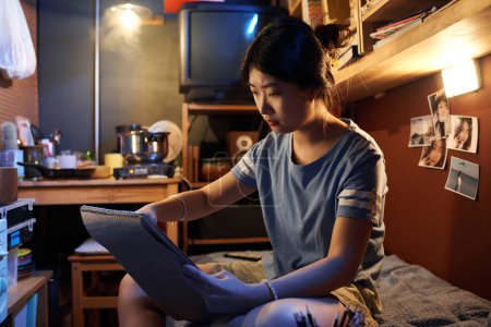 Téléchargez les photos : Fille chinoise assise sur le lit devant la caméra dans un petit appartement et faire un croquis dans un bloc-notes tout en apprenant à dessiner - en image libre de droit