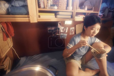 Foto de Zoom asiático femenino sentado en la cama contra estantes de madera colgados en la pared mientras come comida china con palillos para el almuerzo - Imagen libre de derechos