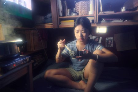 Foto de Mujer asiática joven en pantalones cortos y camiseta sentada en la cama en una habitación pequeña o microplana y comiendo comida china de un tazón con palillos - Imagen libre de derechos
