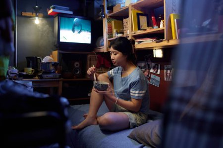 Téléchargez les photos : Jeune femme asiatique avec des baguettes dans un bol de nourriture chinoise assis sur le lit dans un petit appartement, regarder la télévision et déjeuner - en image libre de droit