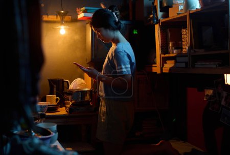 Téléchargez les photos : Image basse clé de jeune femme asiatique avec téléphone portable debout près de la cuisinière électrique dans la cuisine de microflat et préparer le déjeuner - en image libre de droit