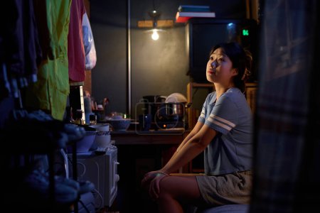 Téléchargez les photos : Jeune femme chinoise en tenue décontractée assise sur le lit dans un petit appartement et regardant des imperméables ou des vestes suspendues à une corde le long du mur - en image libre de droit