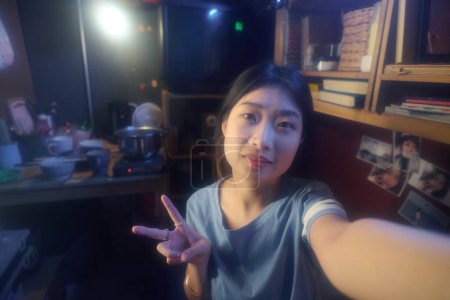 Téléchargez les photos : Heureuse jeune femme chinoise montrant un geste de paix et regardant la caméra tout en prenant des selfies ou en faisant des diffusions en direct de microflat - en image libre de droit