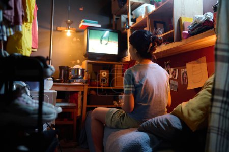 Téléchargez les photos : Vue arrière de la jeune femme asiatique en t-shirt gris assis sur le lit devant l'écran du téléviseur et regardant la diffusion à loisir - en image libre de droit