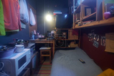 Foto de Pequeño apartamento con mesa de cocina de madera junto al estante con televisor antiguo y otras cosas frente a la cama individual y electrodomésticos - Imagen libre de derechos