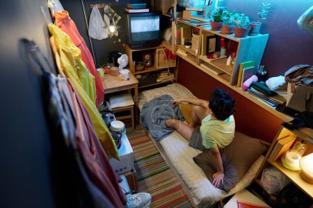 Foto de Alto ángulo de la mujer asiática joven en ropa de casualwear sentado en la cama individual delante de la pantalla del televisor viejo y viendo la emisión o la película - Imagen libre de derechos