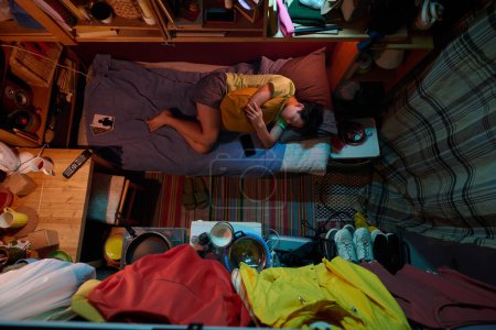 Téléchargez les photos : Au-dessus de l'angle de la fille fatiguée en vêtements de nuit faire la sieste sur un lit simple dans un petit appartement avec cuisine et chambre réunis - en image libre de droit