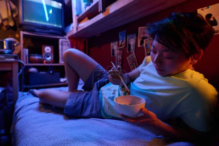 Téléchargez les photos : Fille asiatique affamée avec bol et baguettes relaxant sur un lit simple devant la caméra manger de la nourriture chinoise du bol dans un petit appartement - en image libre de droit