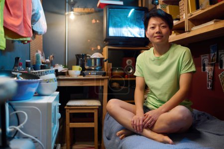 Photo for Young Asian woman in casualwear sitting on bed in small apartment and looking at camera against old TV set and table with kitchenware - Royalty Free Image