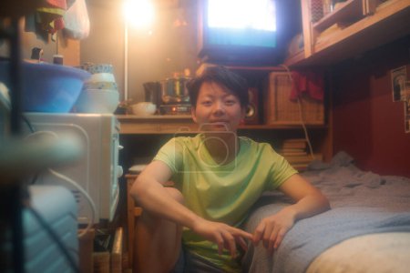 Téléchargez les photos : Heureuse fille asiatique assise sur le sol par un lit simple dans un petit appartement et regardant caméra contre table en bois avec ustensiles de cuisine - en image libre de droit
