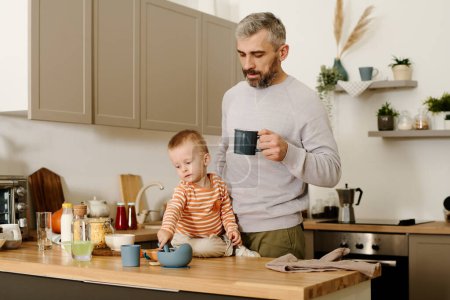 Foto de Padre maduro con taza de té o café de pie junto a su hijo pequeño sentado junto al mostrador de la cocina con utensilios de cocina durante el desayuno por la mañana - Imagen libre de derechos