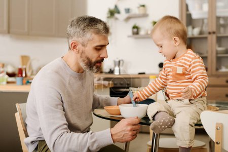 Photo for Mature father sitting by small table in front of his adorable baby son putting spoon into bowl with cornflakes or other food by breakfast - Royalty Free Image