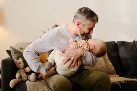 Photo for Mature man in casualwear sitting on couch in front of camera and lulling his cute son after lunch while holding him on hands - Royalty Free Image