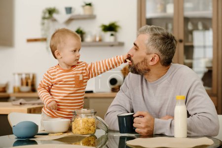 Photo for Adorable baby boy putting cookie or cornflakes into mouth of his father with cup of tea or coffee and enjoying breakfast with his son - Royalty Free Image