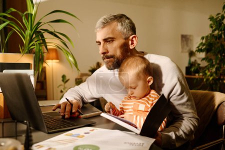 Téléchargez les photos : Homme d'affaires en tenue décontractée regardant l'écran d'ordinateur portable tout en étant assis à table et tenant son mignon bébé fils sur les genoux en regardant la page du livre ouvert - en image libre de droit