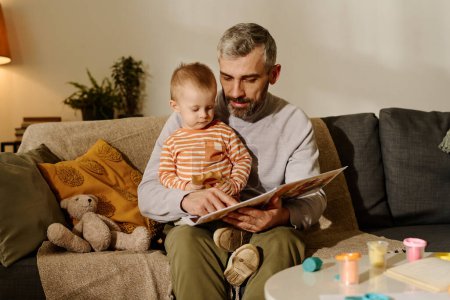 Photo for Mature father in casualwear sitting on couch with his adorable baby son on knees and pointing at page of book while reading fairy tale - Royalty Free Image