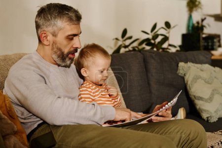 Photo for Man with grey beard and hair pointing at picture on book page and reading it to his adorable baby son while sitting on couch - Royalty Free Image
