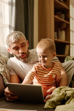 Téléchargez les photos : Mature père et bébé fils regardant l'écran de la tablette tout en se relaxant sur le lit ou le canapé devant la caméra et en regardant la vidéo en ligne ensemble - en image libre de droit