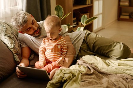 Photo for Mature man and his cute baby son relaxing on bed in front of camera at leisure and watching online video while father looking at infant - Royalty Free Image