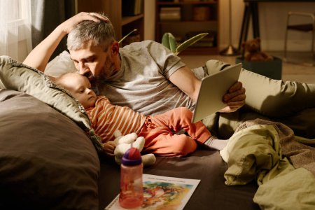 Photo for Man with grey hair kissing his adorable baby son lying next to him and looking at screen of tablet held by mature father - Royalty Free Image