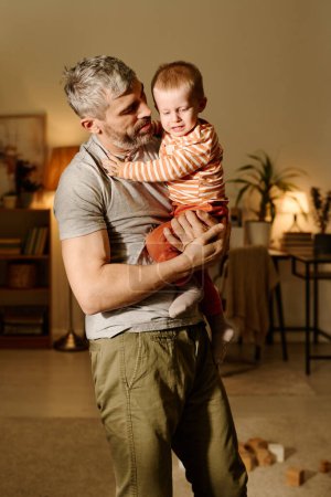 Téléchargez les photos : Homme mûr en t-shirt gris et pantalon beige tenant en pleurs vilain bébé fils sur les mains et le regardant tout en se tenant debout dans le salon - en image libre de droit