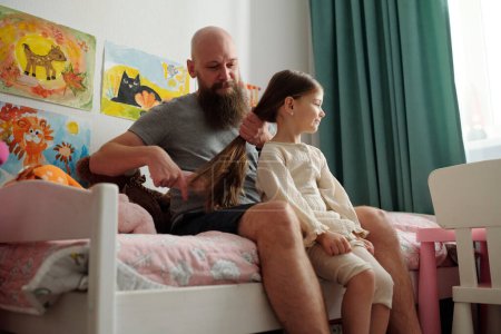 Photo for Young bald man with thick beard sitting on bed and brushing long hair of his adorable daughter against wall with drawings - Royalty Free Image