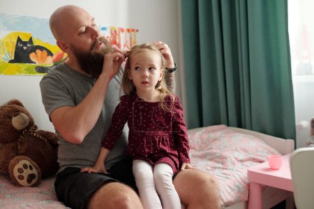Photo for Young bearded man sitting on bed of his adorable blond daughter in smart dress and white tights and braiding her hair - Royalty Free Image