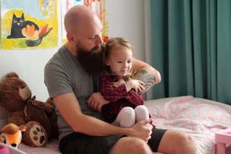 Foto de Joven barbudo con camiseta gris sosteniendo a la linda hija rubia de rodillas y abrazándola mientras ambos están sentados en la cama con juguetes - Imagen libre de derechos