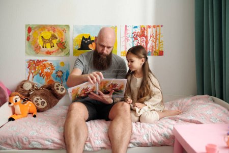 Téléchargez les photos : Homme barbu avec livre ouvert de bandes dessinées avec des photos assis sur le lit à côté de sa petite fille adorable et la lecture de ses nouvelles histoires - en image libre de droit