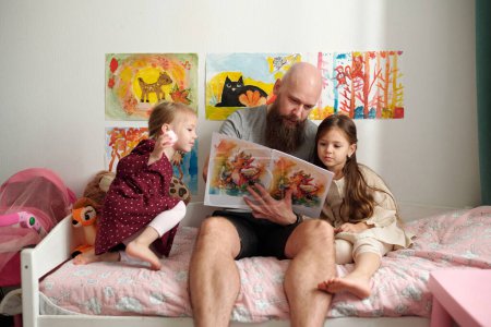 Photo for Bearded man with book of comics showing pictures to one of his cute little daughters while sitting on bed next to girl in white dress - Royalty Free Image