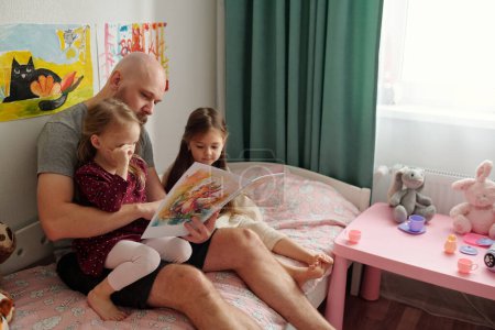 Foto de Barbudo hombre con la cabeza calva lectura libro de cómics con fotos a sus hijas lindas mientras está sentado en la cama entre ellos - Imagen libre de derechos