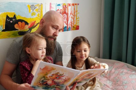 Foto de Joven barbudo discutiendo fotos en libros de cómics con sus dos adorables hijas mientras está sentado en la cama entre ellas - Imagen libre de derechos