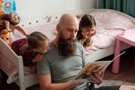 Téléchargez les photos : Père barbu tenant livre ouvert de bandes dessinées tout en étant assis sur le sol par le lit avec ses deux adorables filles regardant des photos - en image libre de droit