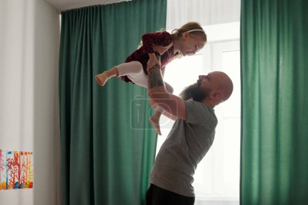 Photo for Happy bearded father holding his cute cheerful daughter on hands while standing in bedroom against green curtains on window - Royalty Free Image