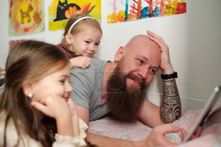 Photo for Happy young man and his two adorable little daughters relaxing on bed in front of camera and watching online movie at leisure - Royalty Free Image