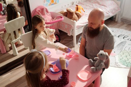 Photo for Young bald man with thick beard sitting by small pink table among his two adorable daughters with toys and having tea with them - Royalty Free Image