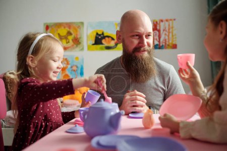 Photo for Happy bearded father looking at one of two adorable daughters during chat while sitting by small pink table and having tea with girls - Royalty Free Image