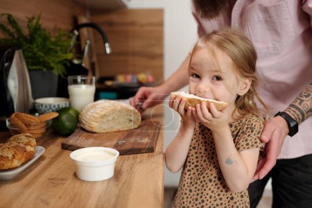 Photo for Hungry little girl eating big sandwich with cream cheese and looking at camera while standing by kitchen counter against her father - Royalty Free Image